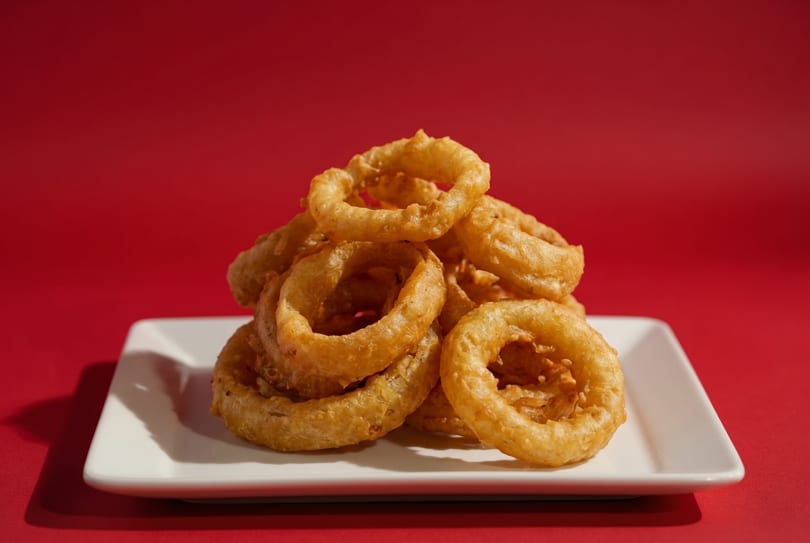 A pile of golden-brown onion rings on a white rectangular plate against a vibrant red background.