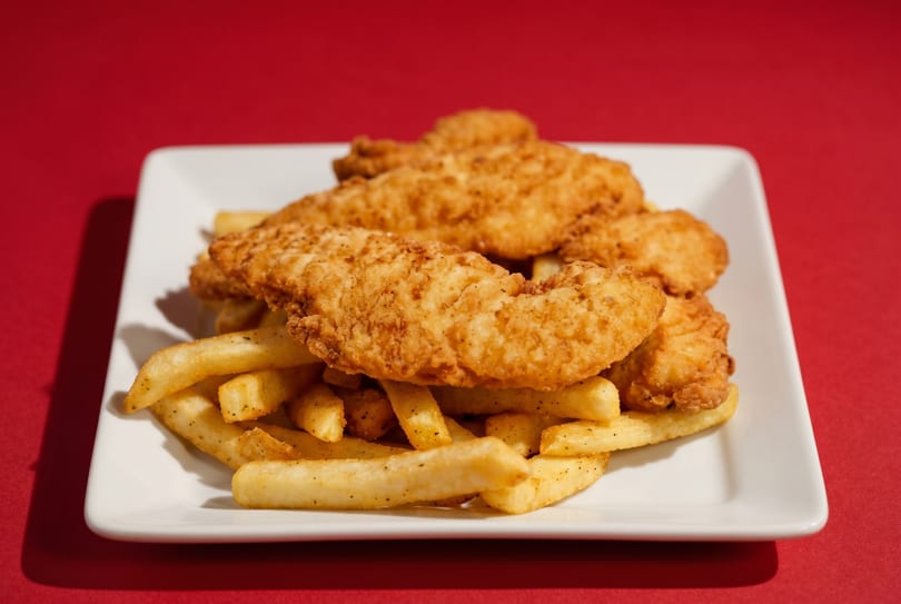 Crispy golden chicken tenders and seasoned french fries on a white rectangular plate against a red background.