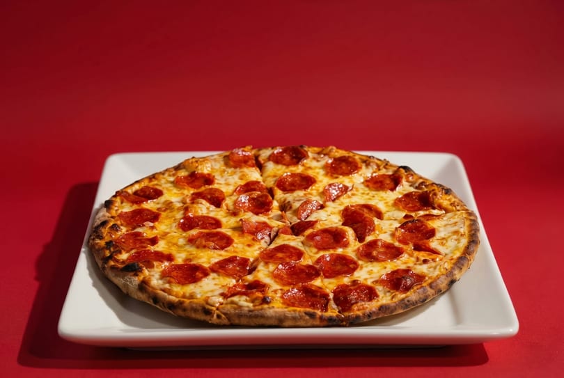 A freshly baked pepperoni pizza, sliced into eight pieces, sits on a white rectangular plate against a red background.