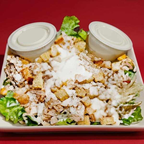 Chicken Caesar salad with croutons and dressing on a white plate against a red background.