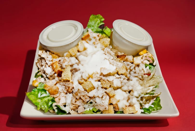 Chicken Caesar salad with croutons and dressing on a white plate against a red background.