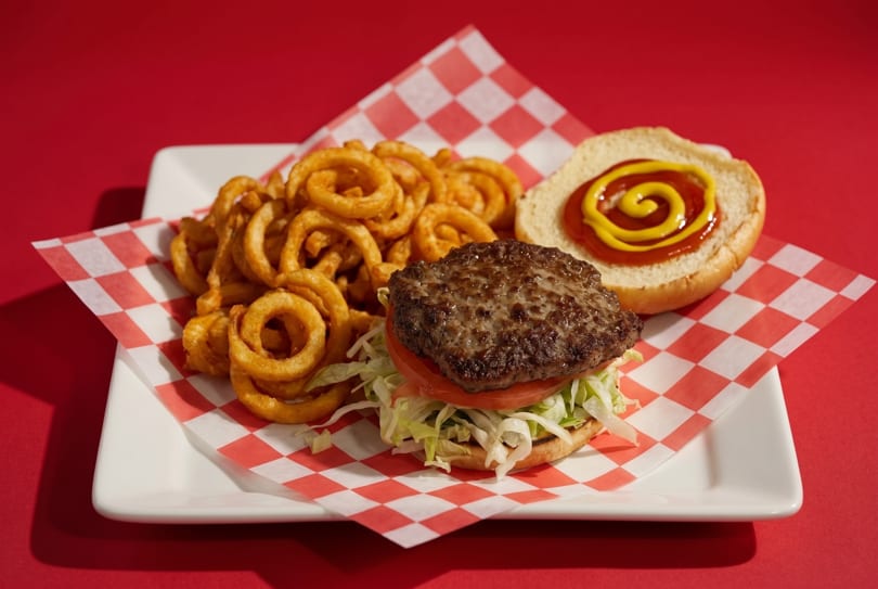 A hamburger with lettuce, tomato, and a side of curly fries on a red and white checkered paper.