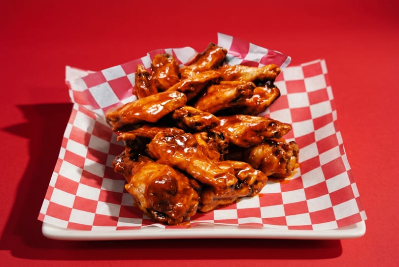 A white plate filled with saucy chicken wings on red and white checkered paper, against a red background.